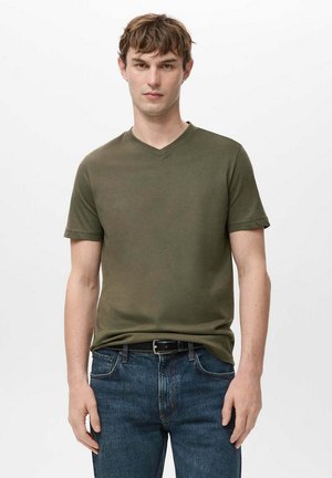 Junger Mann mit kurzen braunen Haaren, der ein olivgrünes V-Ausschnitt-T-Shirt und blaue Jeans mit schwarzem Gürtel trägt, steht vor einem schlichten weißen Hintergrund.