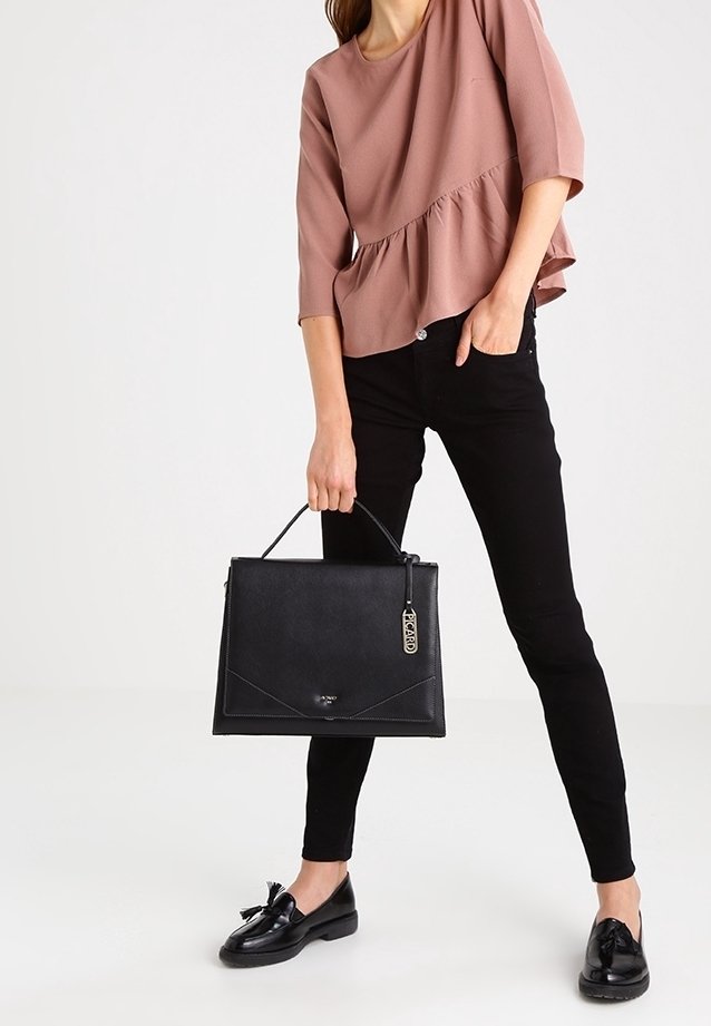 Femme portant un haut mauve et un pantalon noir tient un sac à main noir avec une étiquette, debout en mocassins noirs à pampilles sur fond blanc.