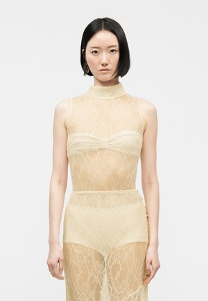 Kvinde med kort sort hår iført en ærmeløs gennemsigtig beige blondekjole med bandeautop og matchende højtaljede shorts, stående mod en hvid baggrund.