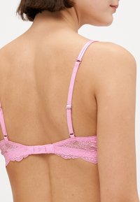 Rosa spetsbh med justerbara band och hak-och-ögla stängning, med en scalloped spetskant längs kanterna. Slät textur.