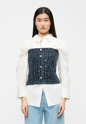 Femme aux cheveux noirs raides portant une chemise blanche à boutons avec de larges manches et un corset sans bretelles texturé bleu marine par-dessus, associé à un jean bleu clair.