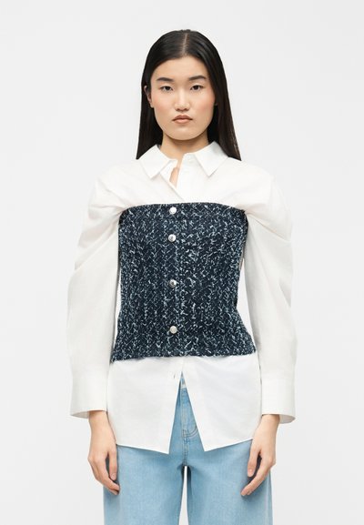 Femme aux cheveux noirs raides portant une chemise blanche à boutons avec de larges manches et un corset sans bretelles texturé bleu marine par-dessus, associé à un jean bleu clair.