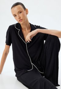 Zwart pyjama set met korte mouwen en witte piping, met een knoopsluiting aan de voorkant en een ontspannen pasvorm. De broek heeft een gestructureerd, geplisseerd ontwerp.