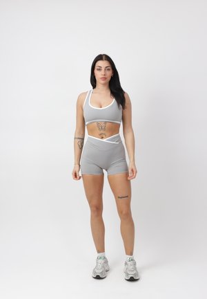 Frau mit langen dunklen Haaren in grauer Sport-BHs und Shorts steht vor weißem Hintergrund und zeigt Schmetterlings- und Schrift-Tattoos.