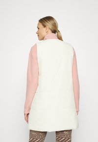 Colete puffer leve e sem mangas em creme, com uma textura suave e design acolchoado. Usado sobre uma blusa rosa e parte de baixo estampada.