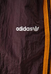 Bordeaux atletisk jacka med en orange mesh-accent och broderad vit Adidas-logotyp. Har en krinklad textur och sidofickdetalj.