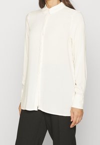 Blusa bianca a maniche lunghe con colletto e patta centrale con bottoni. Tessuto liscio con una vestibilità leggermente sagomata.