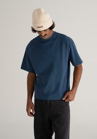 Mies, der ein blaues, Übergröße T-Shirt mit kurzen Ärmeln, schwarze Hosen und eine cremefarbene Strickmütze trägt, schaut nach unten mit einer Hand in der Tasche vor einem schlichten Hintergrund.