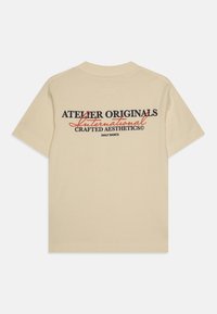 Bež majica iz bombaža z natisnjenim dizajnom na hrbtni strani v mornariško modrih in oranžnih besedah, ki pravi 'ATELIER ORIGINALS International.'