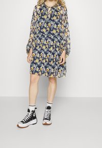 Šaty s květinovým vzorem a dlouhými, nadýchanými rukávy, kulatým výstřihem se zavazováním, v námořnické modré barvě s žlutými a fialovými květy. Černé tenisky.