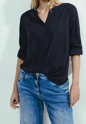 Kvinde iført en mørk navyblå langærmet skjorte med tre knapper og opsmøgede ærmer kombineret med lyseblå denimjeans, stående mod en hvid baggrund.