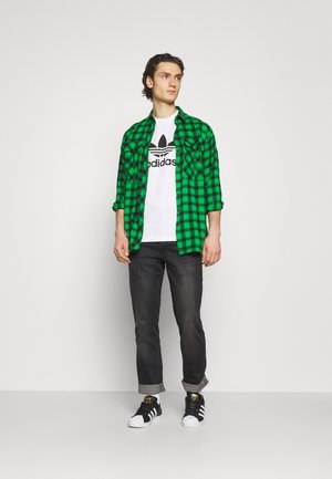 Camisa de franela a cuadros verdes y negros sobre una camiseta blanca con el logo de Adidas, combinada con jeans oscuros y zapatillas negras/blancas.