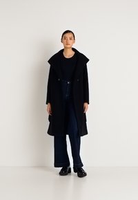 Nő áll egy hosszú fekete kabátban, fekete felsőben, bő szárú sötét farmernadrágban és fekete platform cipőkben egy sima fehér háttér előtt.