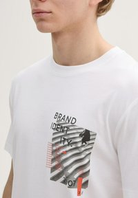 Weißes Baumwoll-T-Shirt mit einem grafischen Druck, der den Text "BRAND IDENTITY", abstrakte Formen und eine schwarze Silhouette auf einem gestreiften Hintergrund zeigt.