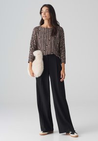 Schwarze weite Hosen kombiniert mit einem gemusterten Oberteil in Brauntönen, hält eine flauschige beigefarbene Handtasche und trägt schwarze Sneaker.