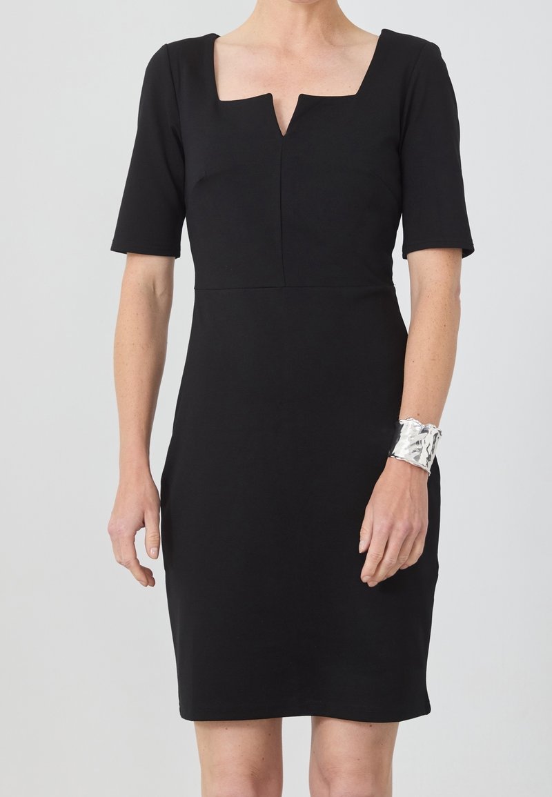 Femme portant une robe noire ajustée avec un décolleté carré en encoche et des manches longueur coude, accessoirisée d’un large bracelet manchette argenté texturé.