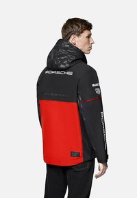 Czarna i czerwona kurtka z wzorzystym kapturem, z logo "Porsche" na plecach i rękawach, wykonana z gładkiego, lekkiego materiału.
