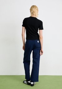 Personne avec des cheveux blonds courts portant un t-shirt noir, un pantalon bleu marine avec un patch logo à l'arrière, et des baskets Adidas noires avec des rayures blanches.