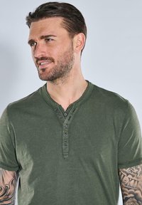Olive green short-sleeve henley shirt with a textured finish, featuring a button placket and a rounded neckline. Tattoo visible on arm.