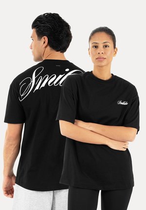 Mężczyzna zwrócony bokiem i kobieta zwrócona przodem, oboje w czarnych koszulkach z białym kursywnym logo „Smile” z przodu i z tyłu.