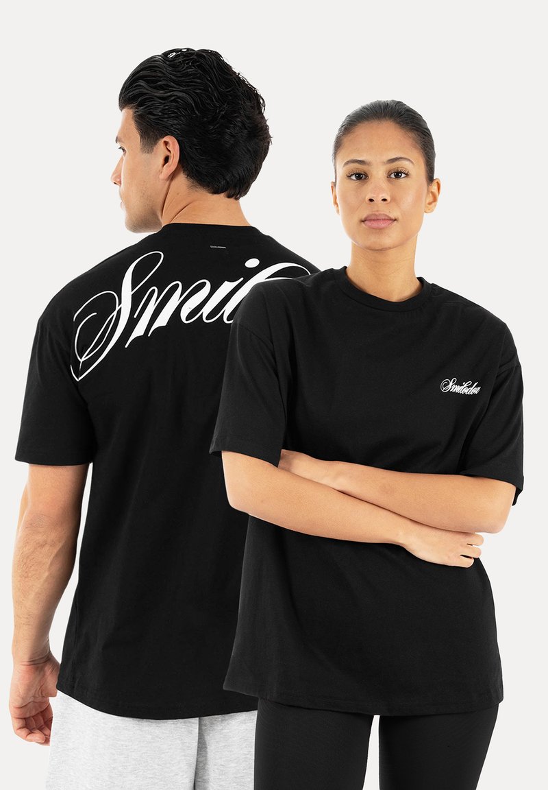 Mann, der seitlich schaut, und Frau, die geradeaus schaut, beide tragen schwarze T-Shirts mit weißen kursiven "Smile"-Logos vorne und hinten.