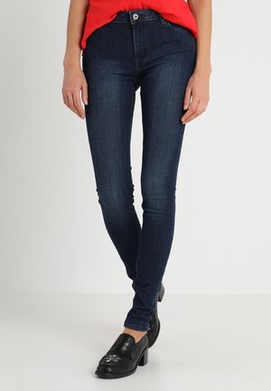 Kvinde iført mørkeblå skinny jeans, rød top og sorte loafers med hæl, stående mod en ensfarvet lys baggrund.