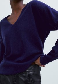 Pull en V bleu marine en tricot doux, avec des manches longues et une coupe décontractée. Associé à un pantalon en cuir noir.
