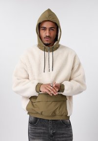 Kremi spalvos megztinis su pašiltinimu, alyvuogių žalia nailono kapišonu ir apatine dalimi. Turi reguliuojamus raištelius bei priekinę kišenę.