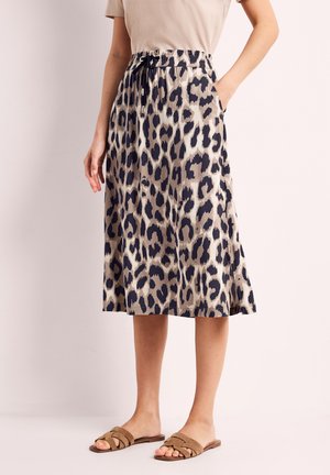 Kvinde iført beige og sort leopardprint midi-nederdel med snøre i taljen, beige top og brune slip-on sandaler, stående med den ene hånd i lommen.