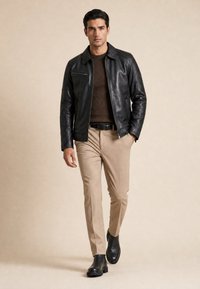 Mann, der vorwärts geht, trägt schwarze Lederjacke, braunen Strickpullover, beige Hose und schwarze Stiefel auf einfarbig beigem Hintergrund.