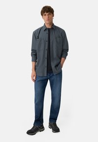 Grijs overhemd met knopen en twee borstzakken, gedragen over een zwart t-shirt. Gecombineerd met blauwe jeans en zwarte sneakers. Neutrale achtergrond.