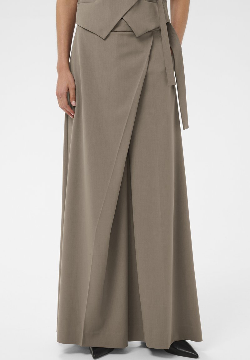 Kvinde iført taube vide omslagsbukser med matchende vest og sorte spidse sko, stående mod en enkel baggrund.