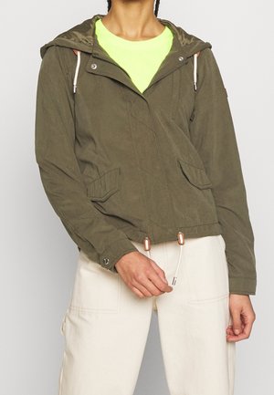 Frau trägt olivgrüne Kapuzenjacke über neongelbem Oberteil und cremeweißer Hose, Hand zieht am Kordelzug der Jacke.