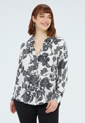 Kobieta uśmiechająca się, ubrana w białą koszulę zapinaną na guziki z czarnym kwiatowym wzorem oraz czarne spodnie, stojąca na jednolitym białym tle.