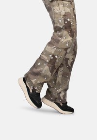 Pantalons cargo camouflage dans des tons de marron et de vert avec des éclaboussures blanches, associés à des baskets noires et beiges avec un design texturé.