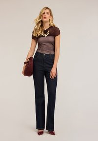 Donna con lunghi capelli biondi indossa un top trasparente marrone, jeans scuri, tacchi bordeaux, una collana dorata e tiene in mano una borsa bordeaux abbinata.