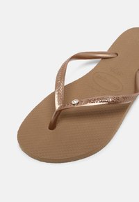 Chinelo castanho com sola de borracha texturizada. Apresenta tiras bronze brilhantes com um design trançado e um pequeno destaque de strass.