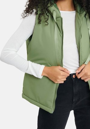 Woman wearing a light green padded vest over a white long-sleeve shirt, fastening black high-waisted jeans.