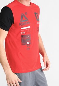 Camiseta deportiva roja con mangas negras, que presenta impresiones gráficas y logotipos en blanco y negro. Textura suave, mangas cortas, diseño de cuello redondo.
