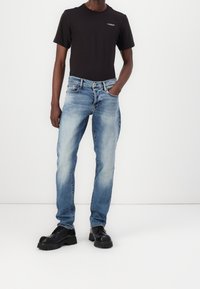 Zwart T-shirt met korte mouwen en een klein logo, gecombineerd met lichtblauwe denim jeans. Model draagt omvangrijke zwarte veterschoenen.
