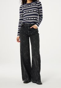 Pruhovaný svetr s dlouhým rukávem v námořnické modré a bílé pletenině s ribbed texturou, zkombinovaný s džínami s vysokým pasem v černé barvě a stříbrnými hroty.