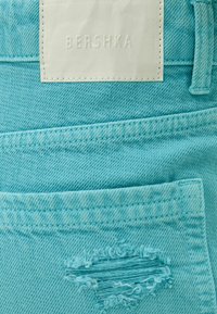 Tela de mezclilla azul claro con un parche desgastado y una etiqueta blanca con relieve que dice "BERSHKA". Costuras visibles a lo largo de los bolsillos y las costuras.