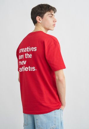 Noor mees punases T-särgis, millel on valge tekst "loojad on uued sportlased" tagaküljel, seljas helesinised teksad, vaatab paremale.