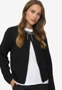 Black ribbed cardigan with an open front and two pockets, featuring a decorative bow tie closure at the neck. Worn over a white shirt.