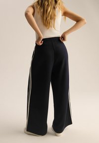 Granatowe spodnie o szerokich nogawkach i luźnym kroju, z białymi paskami po bokach i elastycznym pasem, wykonane z miękkiego, lekkiego materiału.