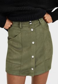 Olivengrüner Wildlederrock mit tailliertem Design, fünf silbernen Druckknöpfen in der Mitte und zwei seitlichen Taschen. Weiche Textur und maßgeschneiderter Look.