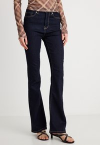 Jeans acampanados de mezclilla azul oscuro con cintura alta, detalles en costura dorada, diseño de cinco bolsillos y acabado suave. Combinados con sandalias negras.