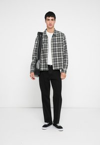 Grå og sort ternet flannelsskjorte kombineret med en hvid t-shirt, sorte bukser og sorte sneakers. Den ene hånd holder en sort taske.