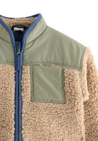 Beige fleecevest met olijfgroene nylonpanelen. Voorzien van een ritssluiting, hoge kraag en een borstzak. Gestructureerd, zacht materiaal.