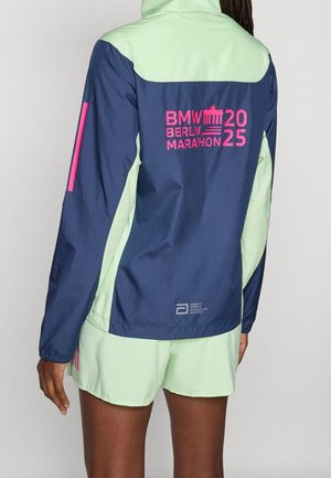 Persoon die een blauw en lichtgroen jack draagt met roze BMW Berlin Marathon 2025-logo en bijpassende lichtgroene korte broek, achteraanzicht.
